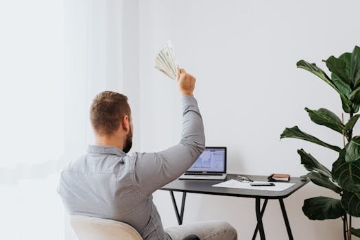 A man in a home office raises cash, symbolizing financial success and achievement. ganhar
