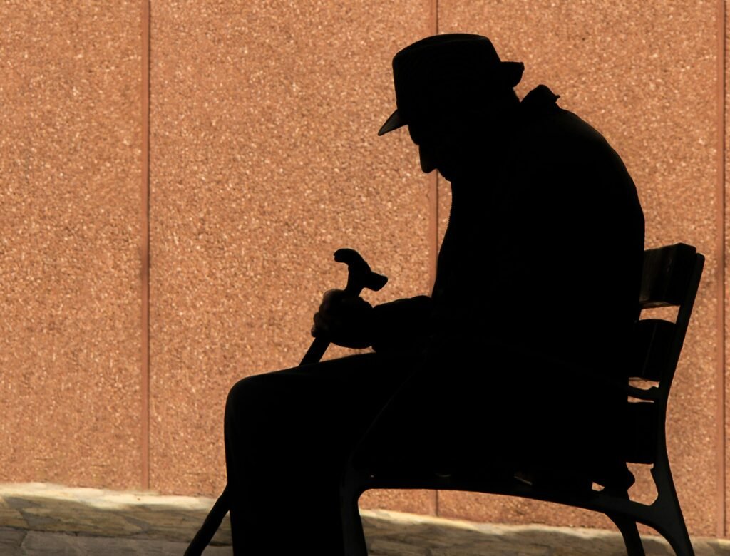 Silhouette of an elderly man with a cane sitting on a bench, capturing a moment of reflection.