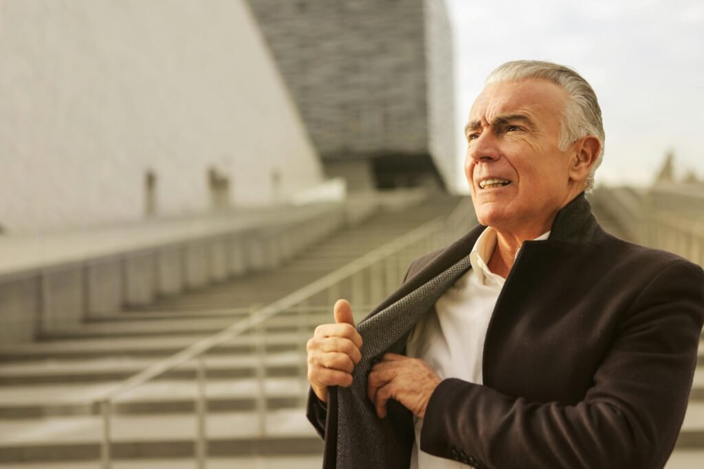 Elderly man confidently adjusting coat, standing outdoors with a modern architectural backdrop.