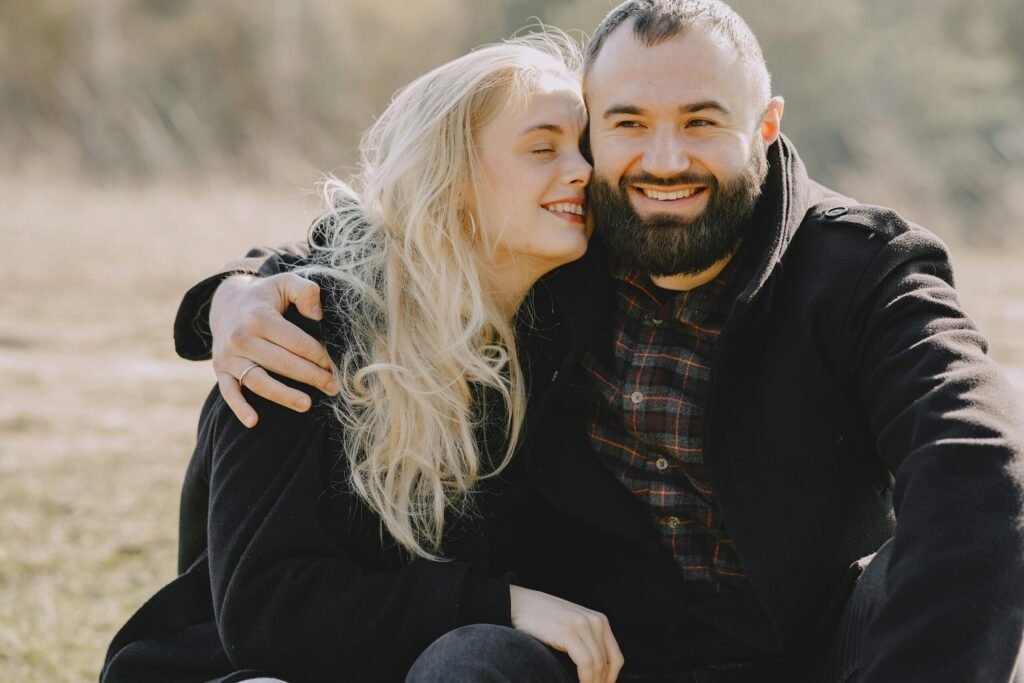 Cheerful lovers in warm wear smiling and embracing each other while spending time together during romantic picnic in park