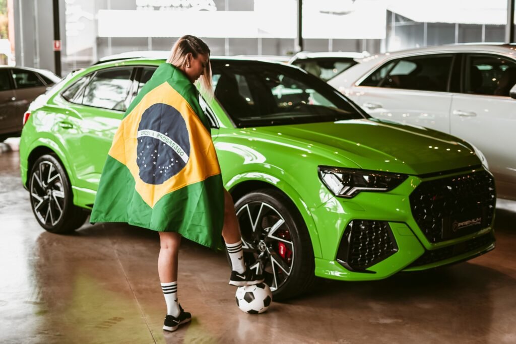 A woman draped in a Brazilian flag with a soccer ball near a green car indoors.
