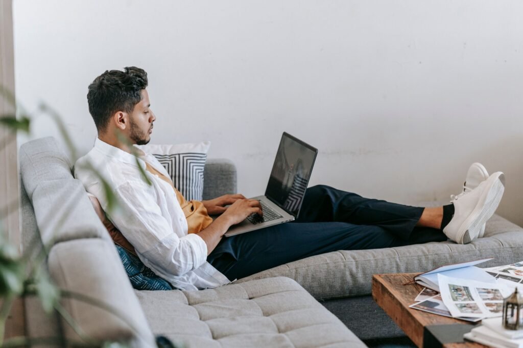 Side view full body of young Hispanic male freelancer sitting on couch and typing on laptop keyboard while working on remote design project at home